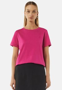 Fuchsia Baumwoll-T-Shirt mit kurzen Ärmeln, Rundhalsausschnitt und dezentem Logodetail. Kombiniert mit einem strukturierten schwarzen Rock.