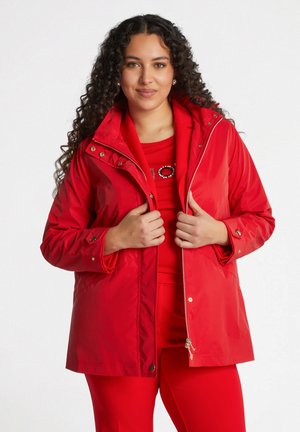 Rode waterdichte jas met ritssluiting, voorzien van een capuchon en drukknopaccenten. Soepele textuur; gecombineerd met een bijpassende rode outfit.