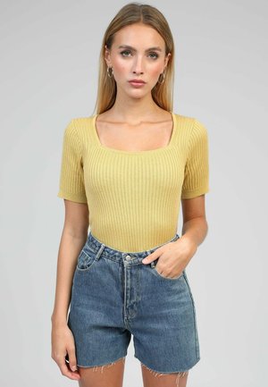 Femme aux cheveux blonds raides portant un haut jaune à manches courtes côtelé et un short en jean bleu taille haute avec un ourlet effiloché.