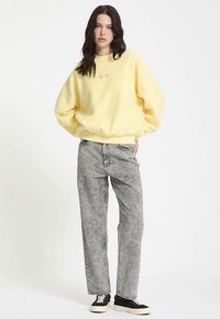 Yellow oversized sweatshirt with embroidered detail, paired with light gray wide-leg jeans and black sneakers with white soles and checkered accents.