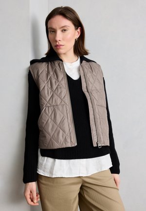 Nő taupe színű steppelt mellényt visel fekete pulóver és fehér ing fölött, bézs nadrággal, egy egyszerű világos fal előtt állva.