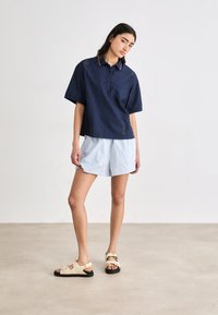 Polo shirt blu navy a maniche corte con dettagli perforati e colletto a contrasto, abbinato a pantaloni corti a righe chiare e sandali beige.