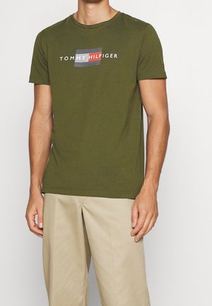 Man draagt een olijfgroene Tommy Hilfiger T-shirt met logo en beige broek, staand tegen een lichte achtergrond.