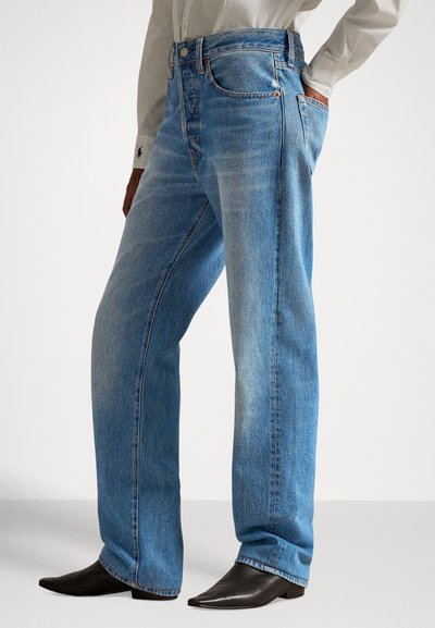 Isik, kes kannab helesiniseid sirge säärega pükse, valget pika varrukaga särki ja musti teravaotstega pahkluu-saapaid, seisab ühe käega taskus.