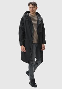 Svart vattentät parka med huva, framzipper och fickor; i kombination med en beige skjorta och grå jeans; elegant, modernt design.