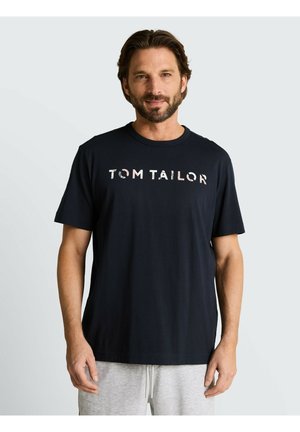 Mężczyzna z krótką brodą i brązowymi włosami, ubrany w czarny t-shirt "Tom Tailor" i jasnoszare spodnie, stojący na jednolitym jasnym tle.