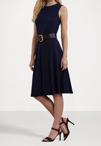Granatowa sukienka bez rękawów, z rozkloszowaną spódnicą i szerokim czarnym paskiem z złotą klamrą. Dobierz do niej czarne szpilki z paskami.