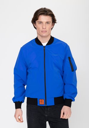 Junger Mann, der eine leuchtend blaue Bomberjacke mit schwarzen Bündchen, Kragen und Reißverschluss trägt, steht vor einem schlichten weißen Hintergrund.