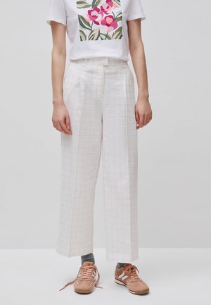 Osoba nosząca białe, szerokie spodnie w czarno-białą kratę z wysokim stanem, białą koszulkę z różowym kwiatowym nadrukiem, szare skarpetki i brązowe trampki.