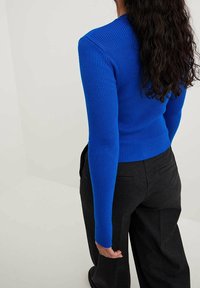 Kobieta o długich, kręconych, ciemnych włosach, ubrana w dopasowany, niebieski prążkowany sweter z długim rękawem oraz ciemnoszare, dopasowane spodnie, stojąca na tle jednolitego tła.