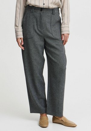 Vrouw draagt grijze broek met hoge taille en brede pijpen met opgestikte zakken, gecombineerd met een beige gestreept hemd en bruine loafers.