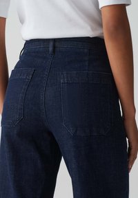 Donkerblauwe denim jeans met een hoge tailleband, twee achterzakken met rechthoekige kleppen en een zachte textuur.