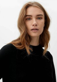 Junge Frau mit welligem, hellbraunem Haar, die einen schwarzen Pullover trägt und leicht an der Kamera vorbeischaut, vor einem schlichten weißen Hintergrund.