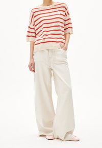 Osoba ubrana w czerwoną i beżową, w paski, bluzkę z krótkimi rękawami, szerokie, kremowe spodnie z troczkiem oraz jasnoróżowe buty, stojąca z jedną ręką w kieszeni.