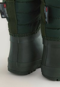 Bottes isolées vertes avec une semelle en caoutchouc texturé, présentant un tissu cousu en haut et des étiquettes indiquant l'isolation et l'étanchéité.