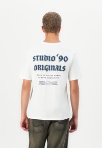 Mann med kort, krøllete hår iført hvit t-skjorte med marineblå tekst "studio '90 originals" og grå bukser, vender bort mot en hvit bakgrunn.