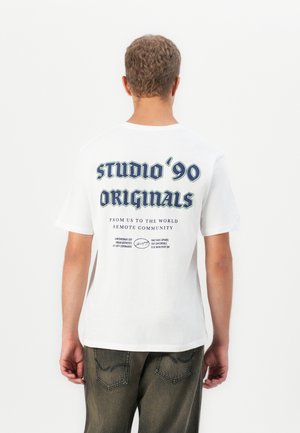 Man with short curly hair wearing white t-shirt with navy text "studio '90 originals" and gray pants, facing away on a white background.
