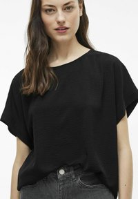 Juodas trumpas rankovėmis viršus su tekstūruotu audiniu, laisvas sijonas ir plati apykaklė, derinamas su aukštu liemeniu pilkais džinsais, kurie turi mygtuko detalę.