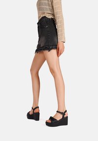 Sandali neri con zeppa e cinturino in pelle annodata, indossati con una gonna di jeans nera con orlo sfrangiato e un top in maglia beige.