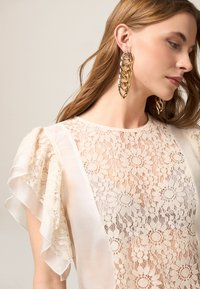 Blouse en dentelle beige avec des manches à volants, ornée de motifs floraux. Assortie à de grandes boucles d'oreilles en chaîne dorée. Texture lisse ; design élégant.