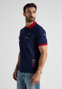 Granatowa koszulka polo z czerwonym kołnierzykiem, krótkimi rękawami, kieszonką na piersi oraz haftowanymi detalami, w tym tekstem i gwiazdkami, po lewej stronie.