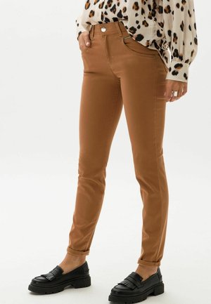 Pantalons en denim bruns ajustés, avec une texture lisse et des ourlets retroussés, associés à des mocassins noirs avec un détail en frange.