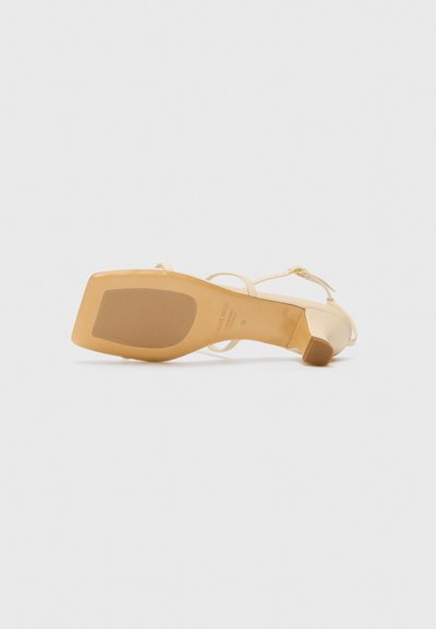 Sandalia de tacón beige con correas finas y punta redondeada. La suela es lisa, con un ligero tacón y detalles de patrón mínimos.