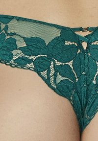String en dentelle teal avec des motifs floraux, présentant une texture en maille transparente et un petit détail en métal au niveau de la taille.