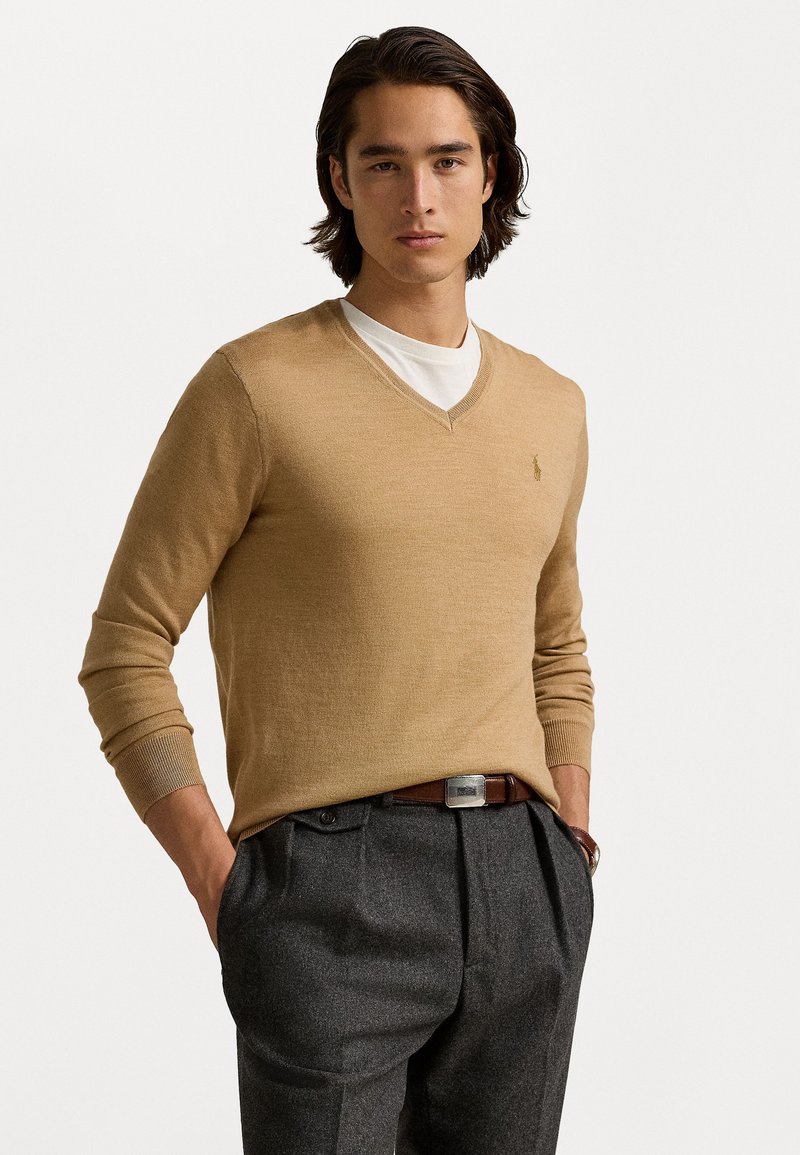 Pull en V beige à manches longues, porté sur une chemise blanche ; tissu tricoté texturé ; associé à un pantalon gris foncé ajusté et une ceinture marron.