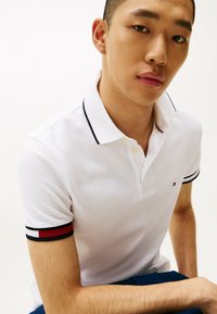 Witte polo met een kraag, voorzien van zwart en rode gestreepte accenten op de mouwen en een klein logo op de borst.