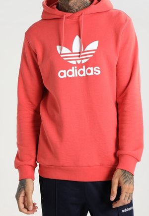 Muž v korálové mikině Adidas s bílým logem trefoil a černých Adidas kalhotách, tetované ruce viditelné na světlém pozadí.