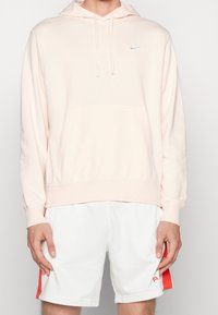 Ljusrosa hoodie med en kenguruficka och dragsko på huvan. Har en liten vit Nike-logga på bröstet. Matchas med vita shorts.