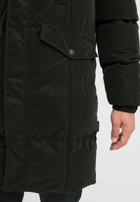 Crna podstavljena parka s teksturiranom površinom, koja ima prednji patentni zatvarač, dva bočna džepa i elastične manžetne. Vidljiva oznaka marke na rubu.