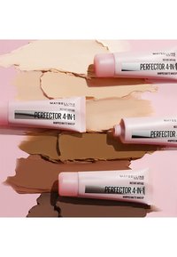 Tubi di trucco opaco montati 4-in-1 di Maybelline in diverse tonalità della pelle su uno sfondo rosa e beige che mette in risalto la texture del prodotto.