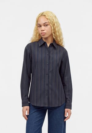 Jeune femme aux longs cheveux blonds bouclés portant une chemise boutonnée à rayures bleu marine foncé et un jean bleu, debout devant un fond blanc uni.