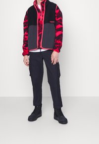 Gilet in pile nero e rosso con zip, caratterizzato da un pannello inferiore nero, abbinato a pantaloni cargo neri e stivali neri.