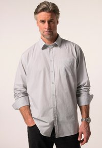 Camicia grigia chiaro con un piccolo motivo geometrico, maniche lunghe arrotolate, taschino sul petto, abbinata a jeans scuri e un orologio da polso.