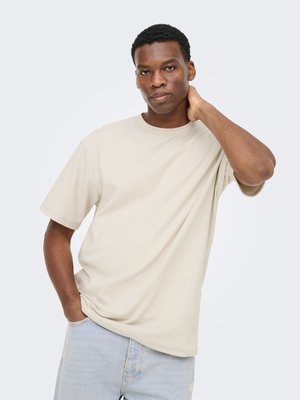 Jonge man met kort krullend haar, een beige geribd T-shirt en lichtblauwe spijkerbroek, hand in zak, andere hand raakt de nek aan, neutrale achtergrond.