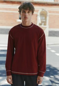 Cămașă burgundy cu mânecă lungă, cu o cusătură albă în contrast în jurul gulerului și manșetelor, purtată cu pantaloni negri. Textură simplă, croială relaxată.