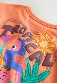 Tissu orange vif avec un oiseau tropical coloré, des feuilles vertes, un soleil cousu et des lettres en relief formant le mot « TROPICAL ».