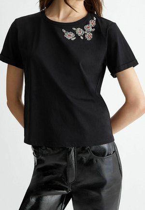 Nainen, jolla on yllään musta lyhythihainen t-paita, jossa helmikirjailtu kukkakuvio lähempänä pääntietä, ja kiiltävät mustat nahkahousut.