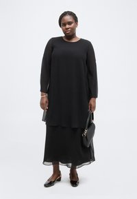 Γυναίκα που φοράει μαύρο φόρεμα με διαφανείς μακριές μανίκες, κρατάει μια μαύρη τσάντα και φοράει μαύρα παπούτσια λουστρίνι με χαμηλό τακούνι, στέκεται μπροστά σε λευκό φόντο.