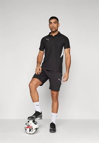 Černé polo tričko a šortky s bílými akcenty, oblečené s černými fotbalovými botami a bílými ponožkami, stojící na fotbalovém míči.