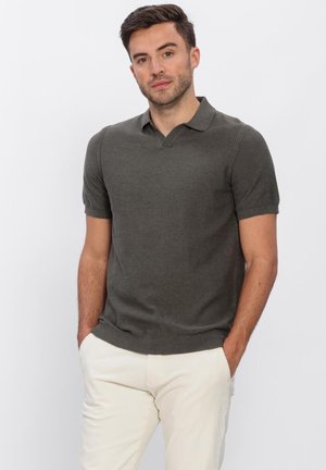 Man staat met handen in de zakken, draagt een donkergrijs polo met korte mouwen en lichtbeige broek tegen een effen achtergrond.