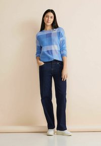 Gemütlicher blauer Pullover mit einem Muster aus hellblauen und dunkelblauen Quadraten, kombiniert mit dunklen, geraden Jeans und weißen Sneakers. Einfaches, lässiges Outfit.