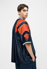 Jaunas vyras su trumpais rudais plaukais, dėvintis mėlynai tamsiai mėlyną ir oranžinį laisvo stiliaus sportinį marškinėlį, stovintis viena ranka kišenėje ir žiūrintis į šoną.