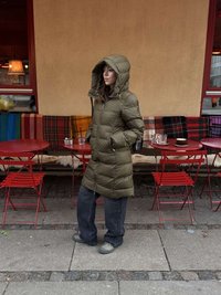 Olivengrøn dunjakke med stor hætte, lynlåslukning foran og synlig polstring. Kombineret med løstsiddende mørke jeans og gråt fodtøj.