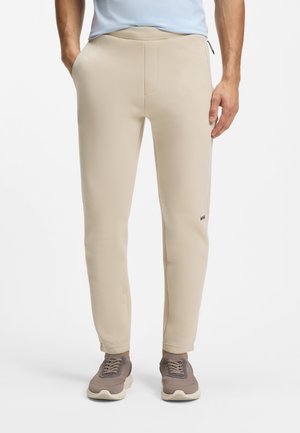 Homme portant un pantalon beige ajusté avec poches latérales et des baskets taupe, debout devant un fond blanc uni.