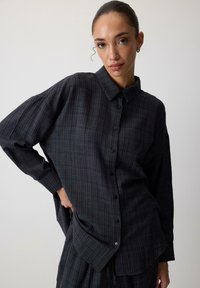 Oversized button-up shirt in dark gray with a checkered pattern, featuring a collar and long sleeves. Made from a textured fabric.