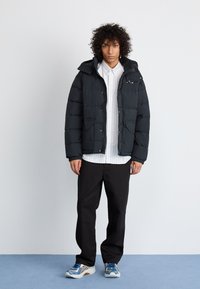 Schwarze Pufferjacke mit Kapuze, Reißverschluss und zwei vorderen Taschen. Getragen über einem gestreiften weißen Hemd und kombiniert mit schwarzen Hosen und blauen Turnschuhen.